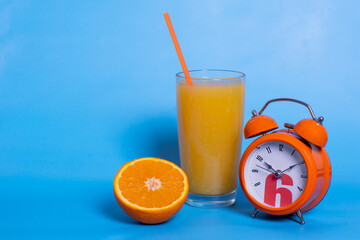 Fresh orange juice in a glass, fruit cut in half, orange alarm clock isolated on a blue background, copy space