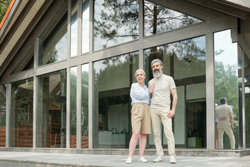 Content stylish senior couple embracing against glass cottage while waiting for guests outdoors