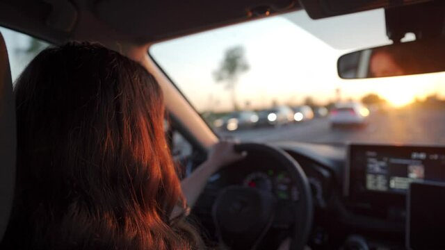 Beautiful brunette woman with long hair driving car at sunset sun rays shine through car windshield rear view of adult woman drives automobile on highway at dusk.
