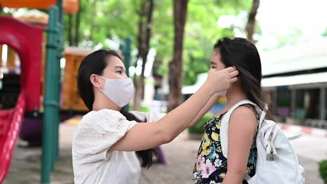 Mother puts a safety mask on daughter face for protection Covid-19 or coronavirus outbreak in village park to prepare go to school. Back to school concept. Medical mask to prevent coronavirus.