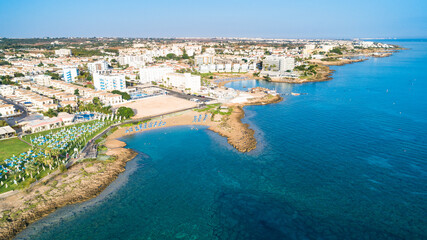 Fototapeta premium Aerial bird's eye view of Pernera beach in Protaras, Paralimni, Famagusta, Cyprus. Tourist attraction golden sandy bay with sunbeds, water sports, hotels, restaurant, people swimming in sea from above
