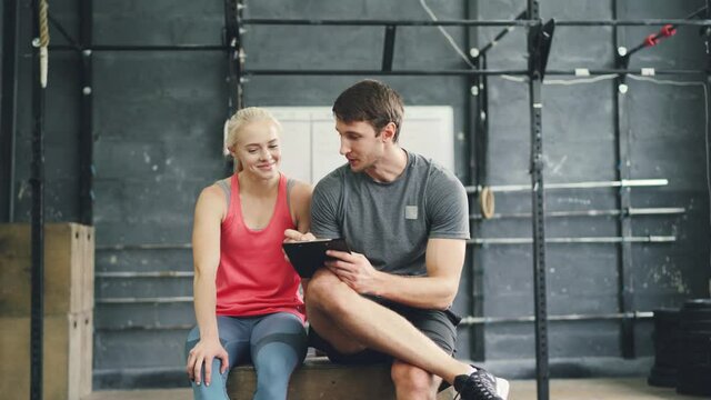 Joyful girl and guy are talking relaxing after training in crosfit gym, trainer is writing in chart planning physical activity. People, communication and sports concept.