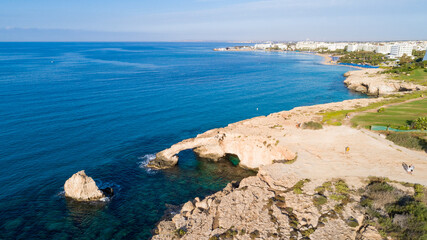 Aerial bird's eye view of coastline landmark ‘Love bridge’ and sea caves at Cavo Greco, Ayia Napa, Famagusta, Cyprus from above. A tourist attraction cliff rock natural formation arch in Ammochostos.