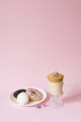 Beautiful dalgon coffee drink. A Cup of coffee with foam and lilac flower, a plate of sweets: marshmallows and donuts on a pink background.