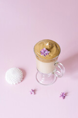 Beautiful dalgon coffee drink. Coffee mug in clear glass with foam and lilac flower on a lilac background. Sweetness to the drink-marshmallow