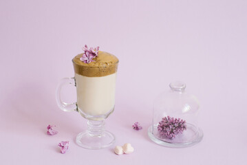 trendy drink 2020 dalgon Coffee in a transparent Cup with a paper tube, a plate with lilac flowers on a lilac background with a copy space
