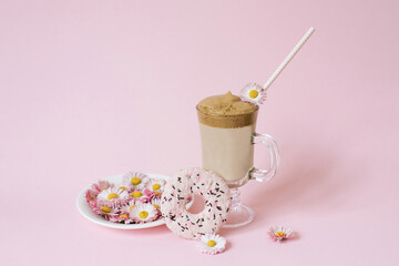 dalgona Coffee in a transparent Cup with a paper tube, a plate with flowers daisies and a delicious mouth-watering Donat with pink icing