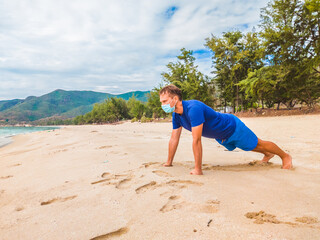 Coronavirus COVID 19. Young beautiful man doing sport performing workouts exercises and push up near sea in the morning during quarantine. Active life in surgical sterilizing face mask protection