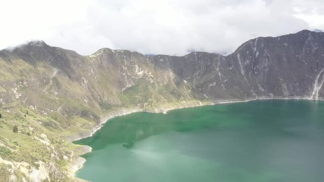 Aerial view of the Quilotoa lake in the crater of an inactive vulcano while the sun is shining down showing a green blue coloration in the water
