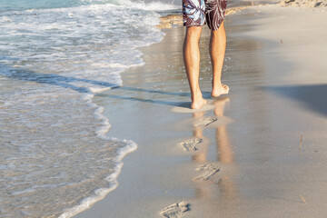 Male feet on the shore of a sandy beach on a sunny day. A young man walks along the edge of the water leaving footprints. Space for text. Tourism and rest.