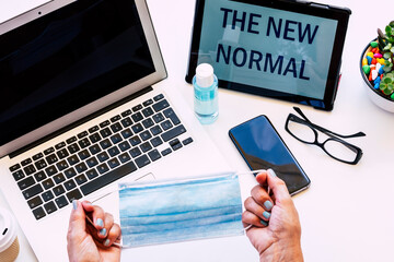 Senior woman holding a facial mask with hands - white desk with laptop computer and tablet with message - the new normal