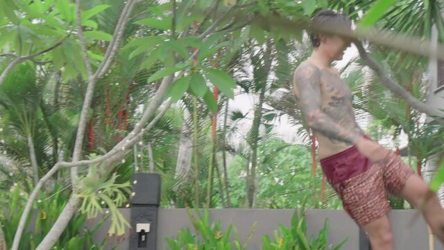 Young man with tattoo on his body and wet hair wearing shorts riding skateboard on halfpipe in villa backyard, Bali
