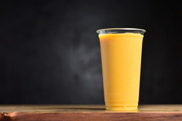 Fruit smoothie in a plastic glass. Summer drink of mango, banana, ice and juice on a black background with copy space