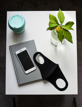Photo from the top of a reusable face mask, an electronic device a coffee and a plant on a white table