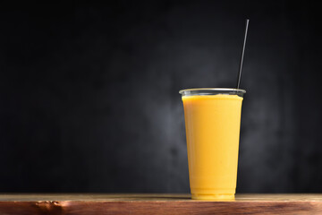 Fruit cocktail in a plastic glass and a straw tube. Summer drink of mango, banana, ice and juice on a black background with copy space