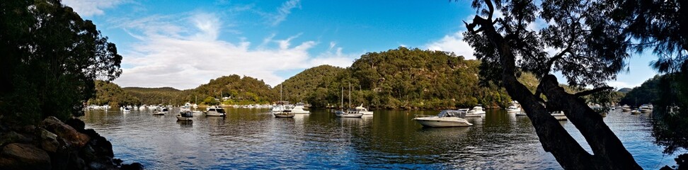 Beautiful morning panoramic view of Creek with boats and reflections of blue sky, light clouds, mountains and trees. Berowra Waters, Berowra Valley National Park, New South Wales, Australia