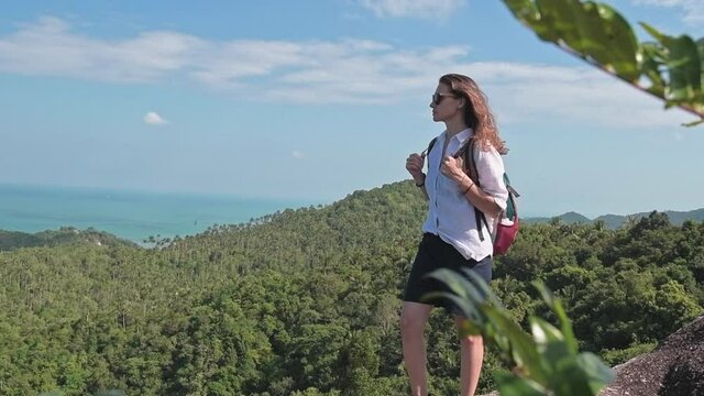 A young, beautiful, active woman with a backpack stands on top of a mountain. A cute girl stands on the edge of a cliff overlooking the blue sea. The wind develops the hair of a tourist.