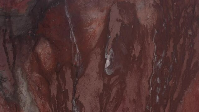 Still video showing a relatively small waterfall of meltwater from a large gletser surrounded by red vulcanic stones
