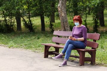 Aged, matured woman with red hair in a medical mask on a bench in a park, uses phone..