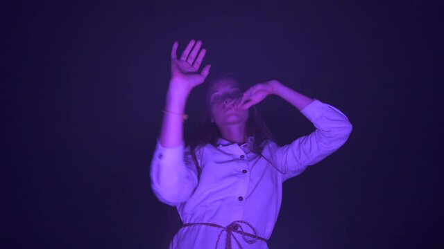 Slow ecstatic dance of young beautiful long haired woman in white dress in the darkness. Purple light. Low angle shot. Night club dancing party. Musical mood.
