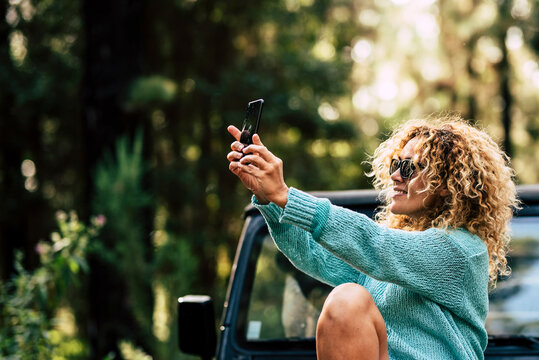 Outdoor people enjoy technology with cheerful caucasian woman take a picture with smartphone in the forest during a travel with her car - adult female and alternative vacation with nature