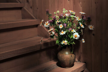 Fototapeta na wymiar Rustic still life with daisies in an old clay jug on a wooden staircase in a village house.