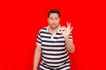 Brunette young man in striped t-shirt showing ok sign isolated over red background