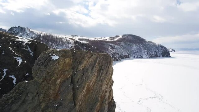 Steep rock and the shore of a frozen lake. Clip. Aerial view of the steep cliff and mountains covered by bald trees and the lake under the ice and snow, winter nature landscape.