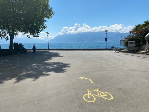 Asphaltierte Promenade am See. Vorne ein Fahradsymbol auf der Stra&szlig;e mit Pfeil. Baum mit mensch als Shilouette. Hinten der See, Berge mit wei&szlig;en Wolken umrandet.