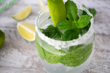 Mojito refreshing cocktail drink with ice, lime and mint in a glass on a light wooden background. Lime slices lie near. The edges of the glass are grated with lime and covered with salt, close-up.