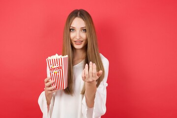 Young caucasian woman holding bucket with popcorn standing over isolated red background inviting to come with hand. Happy that you came