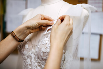 Close-up of work process of a tailor in her studio. Hands sewing bridal dress process, pinning lace on mannequin.
 - obrazy, fototapety, plakaty