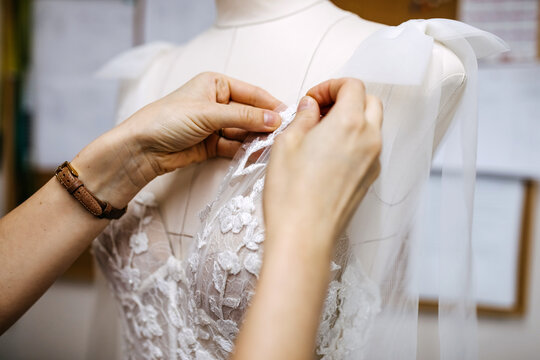 Close-up of work process of a tailor in her studio. Hands sewing bridal dress process, pinning lace on mannequin.

