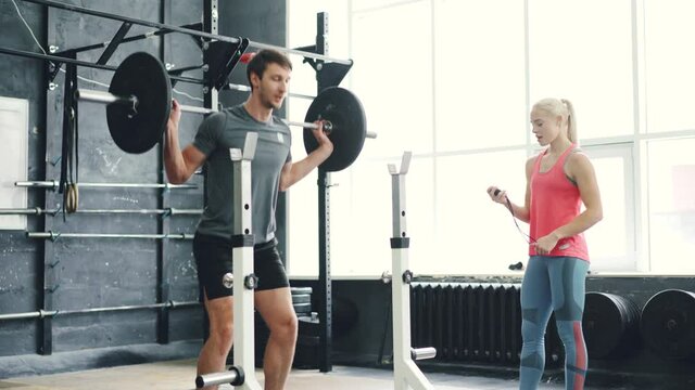 Guy in sportswear is squatting with barbell in gym training body working out with female coach indoors. Physical activity, people and modern lifestyle concept.