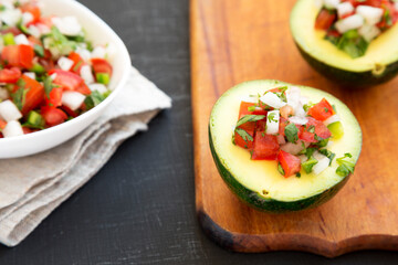 Homemade Pico de Gallo Stuffed Avocado on a rustic wooden board on a black background, side view. Close-up.