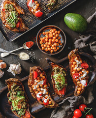 Baked sweet potato toasts with roasted chickpeas, tomatoes, goat cheese, avocado, seedlings on wooden board over brown background. Healthy vegan food, clean eating, toning, top view