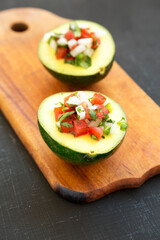 Homemade Pico de Gallo Stuffed Avocado on a rustic wooden board on a black background, side view. Close-up.