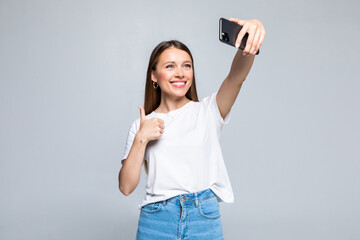 Fototapeta premium Happy cheerful young woman showing thumb up and making selfie on smartphone isolated on white background