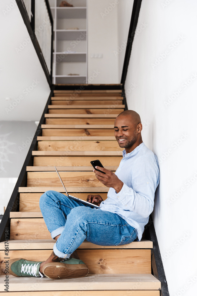 Wall mural Photo of smiling man using laptop and cellphone while sitting on stair - Wall murals
