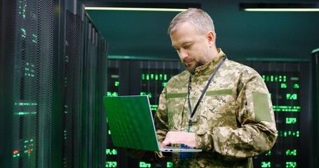 US army senior Caucasian male soldier working at laptop computer in dark room with monitors....