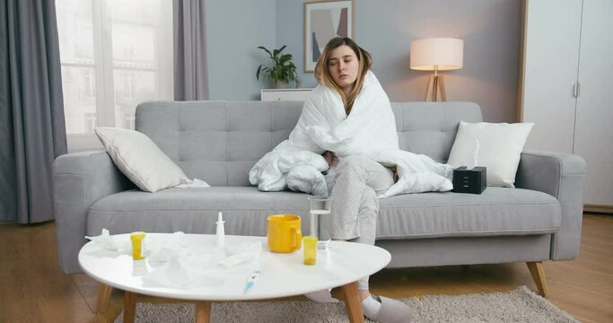 Tired unhealthy young caucasian woman sitting on couch in living room under blanket with high temperature and fever. Ill female caughing and blowing nose in napkin at home. Girl on sofa with flu.