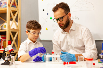 dad and son the child stayed at home conduct experiments on a white table, behind them on the wall is a white Board magnetic-marker