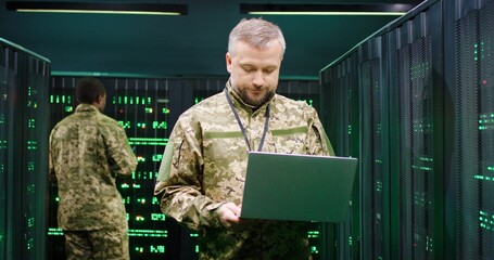 Handsome Caucasian military cybersecurity worker typing on laptop computer while standing in server...