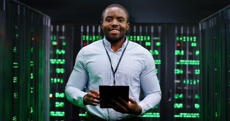 Portrait of young handsome African American male big data specialist wih tablet device in hands smiling to camera in servers room. Happy man in database center. Digital storage concept.