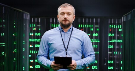 Portrait of handsome Caucasian male big data specialist wih tablet device in hands looking at camera in servers room. Businessman in database center. Digital storage concept. Telecommunication.