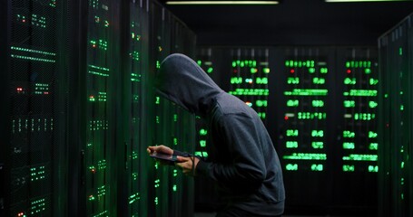 Male Asian guy data thief in dark hood stealing in the server room with tablet device in hands....
