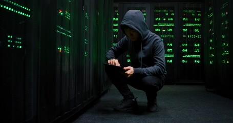 Masked hacker in hood sitting at big server with personal data storage and breaking into system while searching for passwords on tablet device. Database thief stealing information.