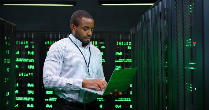 Male African American big data analytic walking at server dark room, working with secret data and using laptop computer to check the information. Monitoring database center. Cloud storage of info.