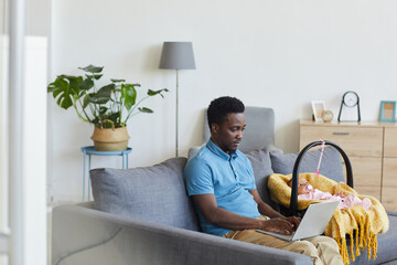 African man working online on his laptop while sitting on sofa with his baby