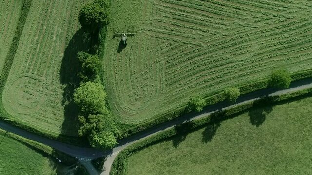 Top down of a rural setting. A country road T junction, and in the field next a farmer in his tractor tends to his cut grass field